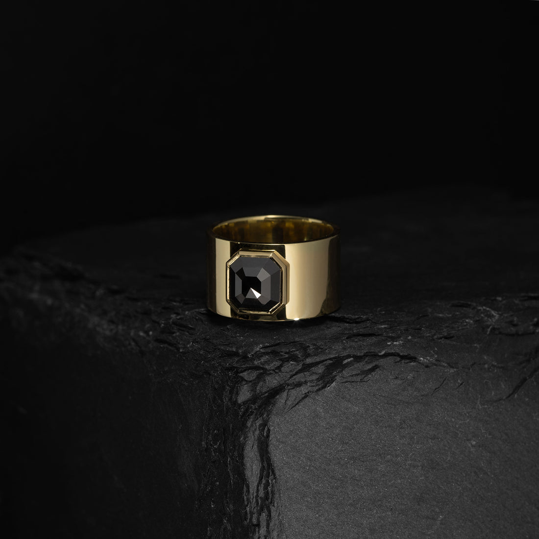 ILA SODHANI Aubergine Black Ring featuring a black cushion cut spinel stone set into a wide yellow gold band; shown on black stone in front of a black background.