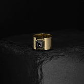 ILA SODHANI Aubergine Black Ring featuring a black cushion cut spinel stone set into a wide yellow gold band; shown on black stone in front of a black background.