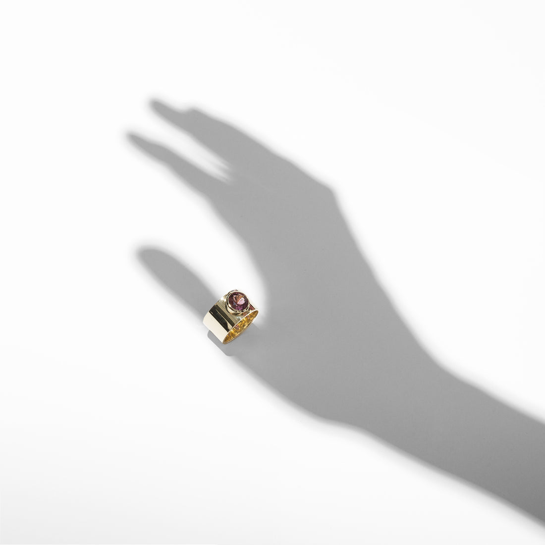 ILA SODHANI Aubergine ring with a magenta natural spinel stone on a wide yellow gold cigar band; shown with a shadow positioned to look like the ring is worn on the thumb.