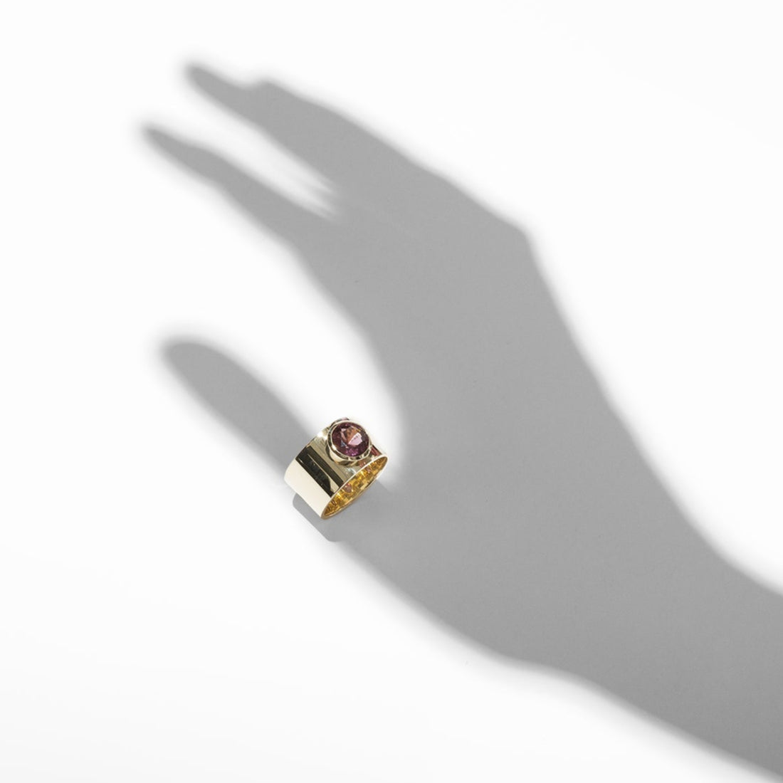 ILA SODHANI Aubergine ring with a magenta natural spinel stone on a wide yellow gold cigar band; shown with a shadow positioned to look like the ring is worn on the thumb.