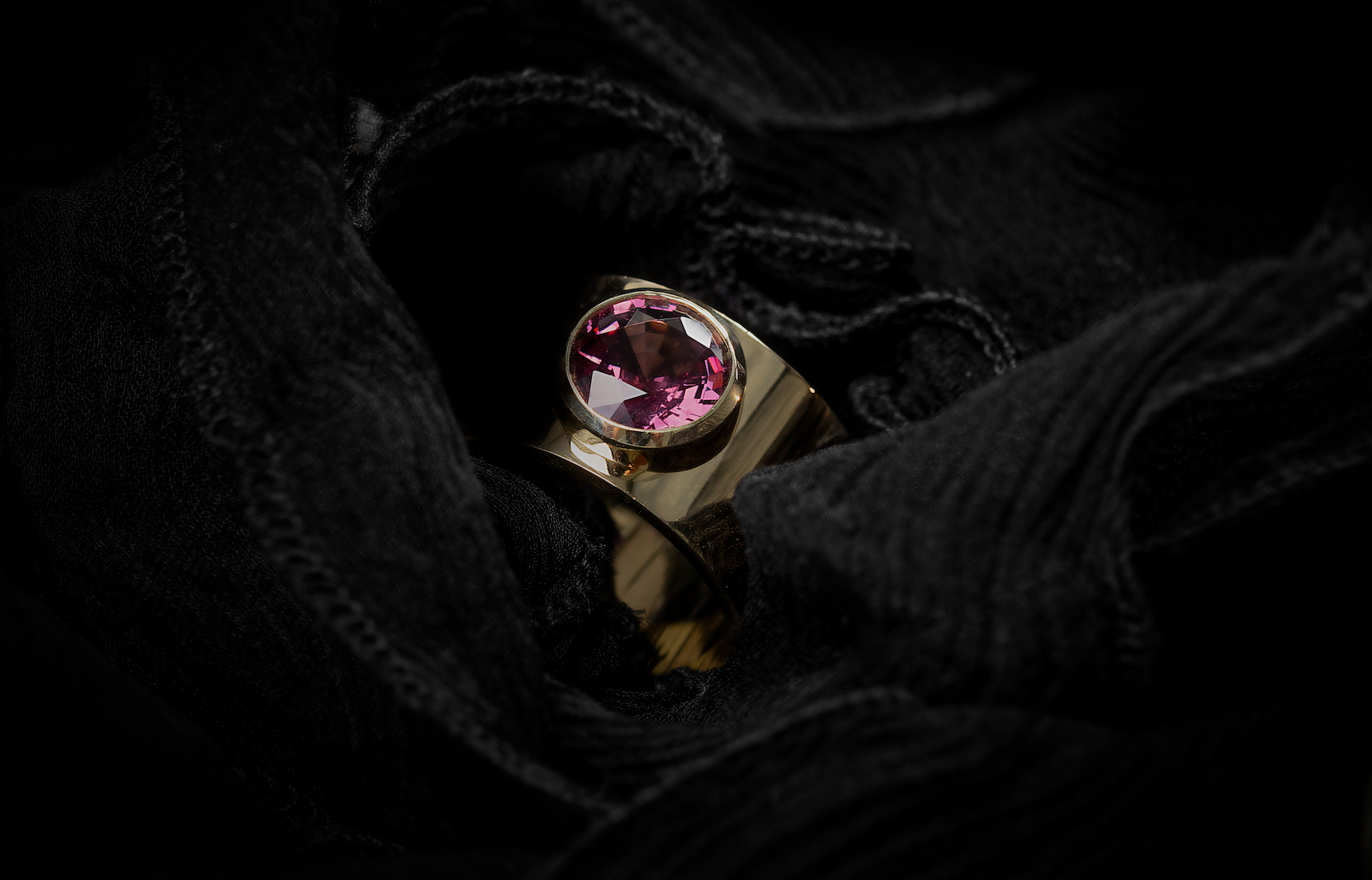 ILA SODHANI Aubergine Ring with a magenta natural spinel stone set into a wide yellow gold band; shown nestled amongst black fabric.