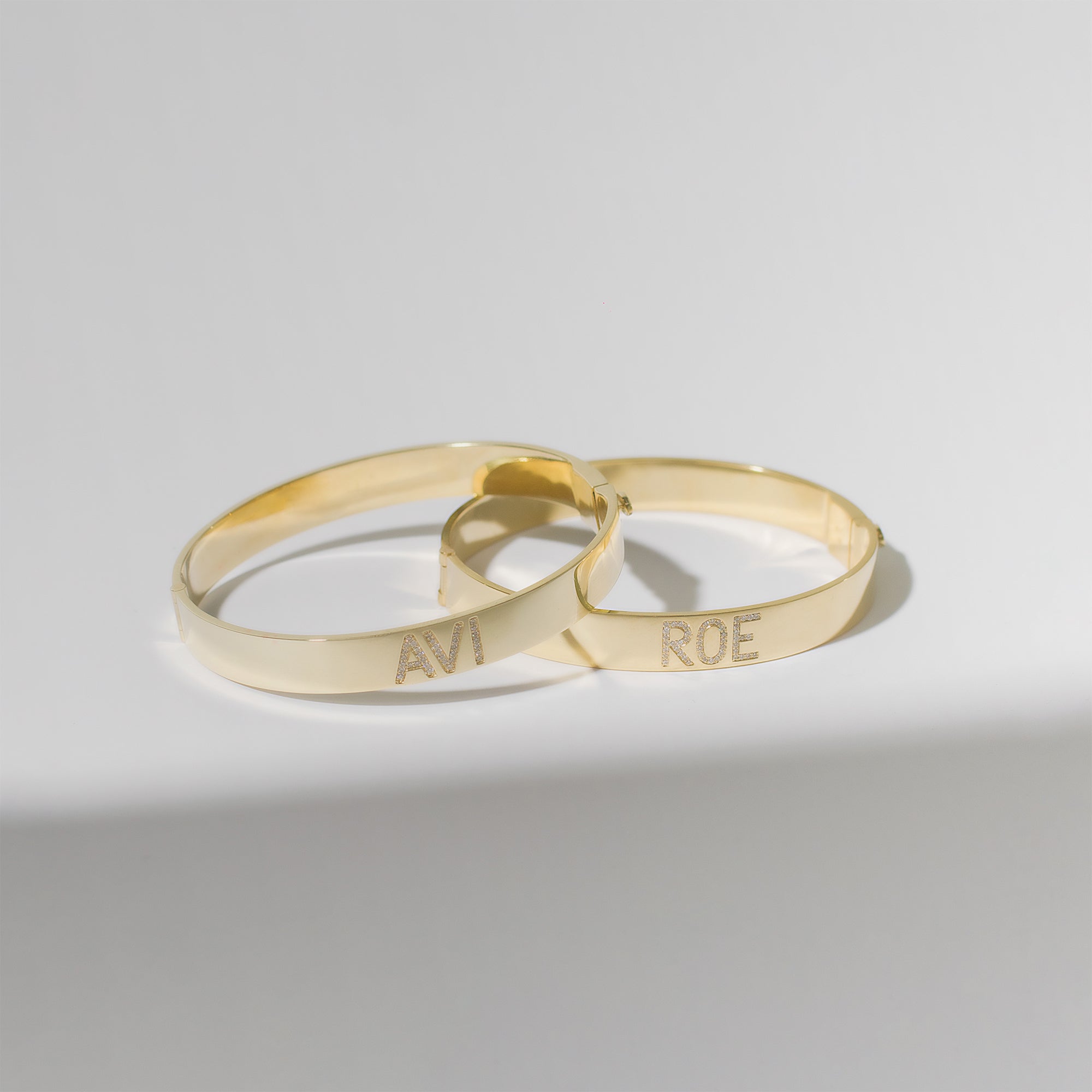 Two of ILA SODHANI's Celebration Bangles in yellow gold with diamonds set into the names "Avi" and "Roe"; the bangles are leaning on each other in front of a white background.