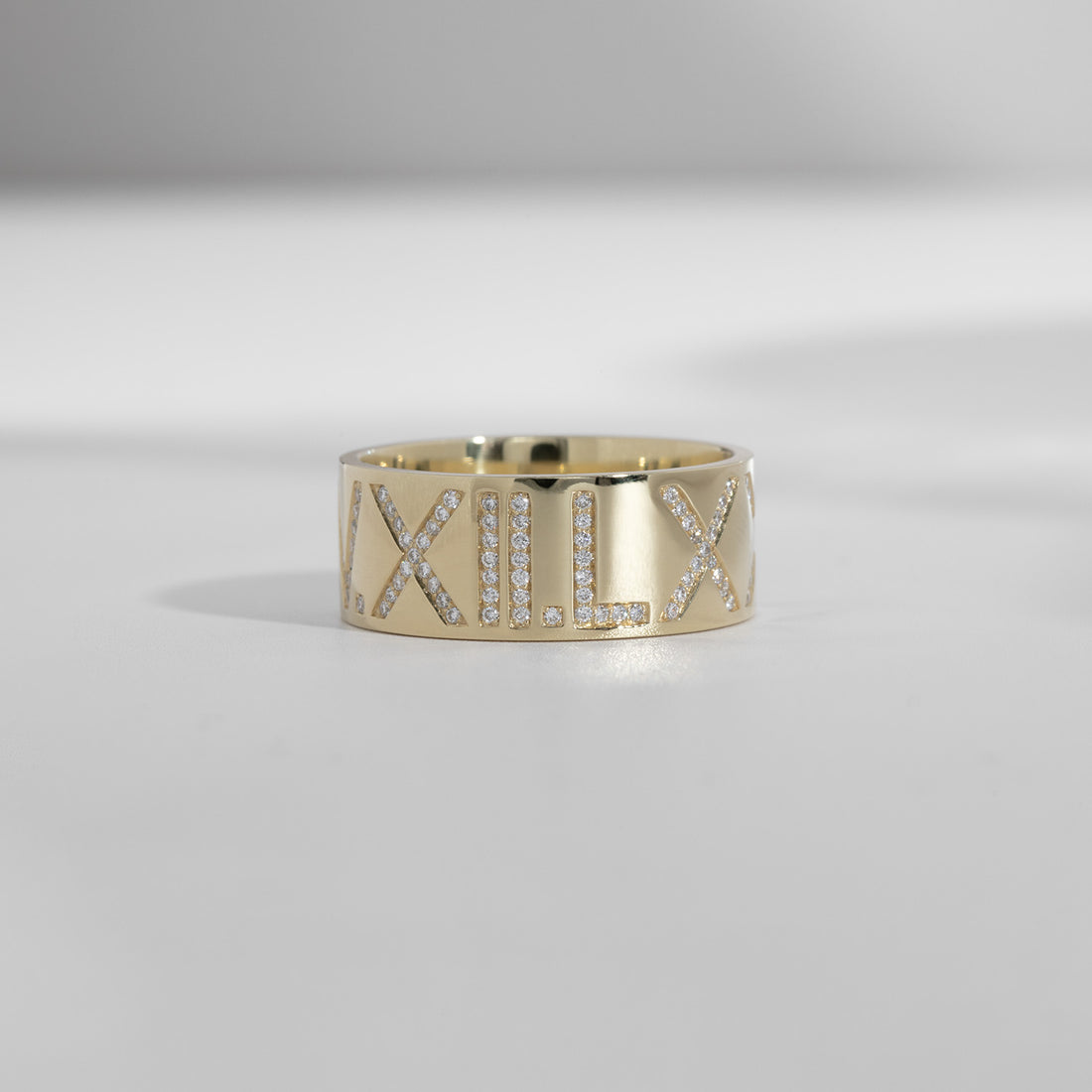 An ILA SODHANI Celebration Ring in yellow gold with white diamonds set into the roman numerals, &quot;V.XII.LX&quot;; shown in front of a white background.