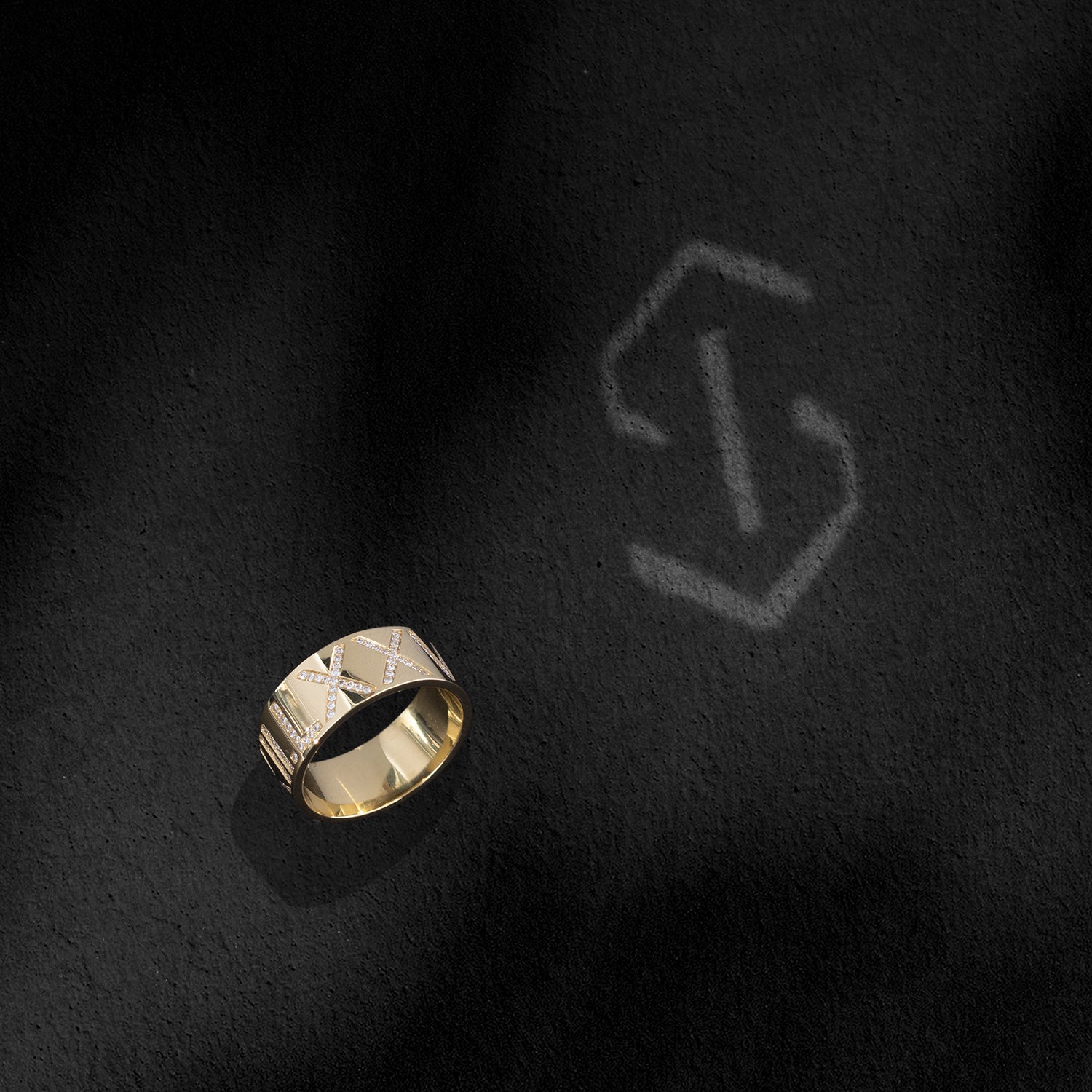 An ILA SODHANI Celebration Ring in yellow gold with white diamonds set into the roman numerals, &quot;V.XII.LX&quot;; shown standing next to the ILA SODHANI logo cast in light onto a of a black background.