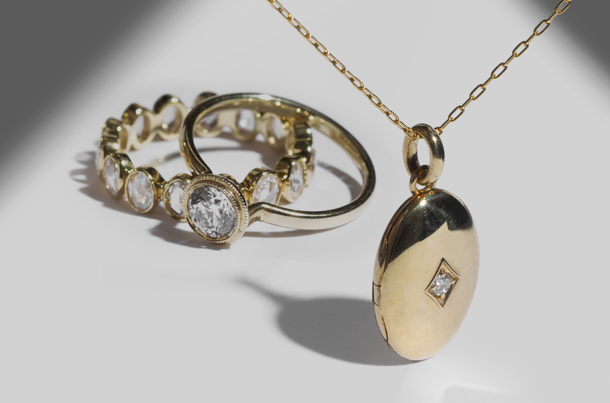 ILA SODHANI's Ellipse Band and Bezel Diamond Engagement Ring in yellow gold stacked behind the Tara Locket in yellow gold hanging in the foreground; shown on a white background with shadows in the upper corners.