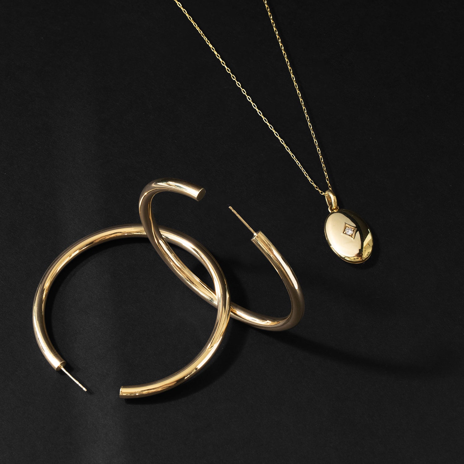 ILA SODHANI's Medium Addax Hoops in yellow gold intertwined, shown with the ILA SODHANI Tara Locket in yellow gold with an oval pendant that has a natural diamond set in an etched diamond space, on a black background.