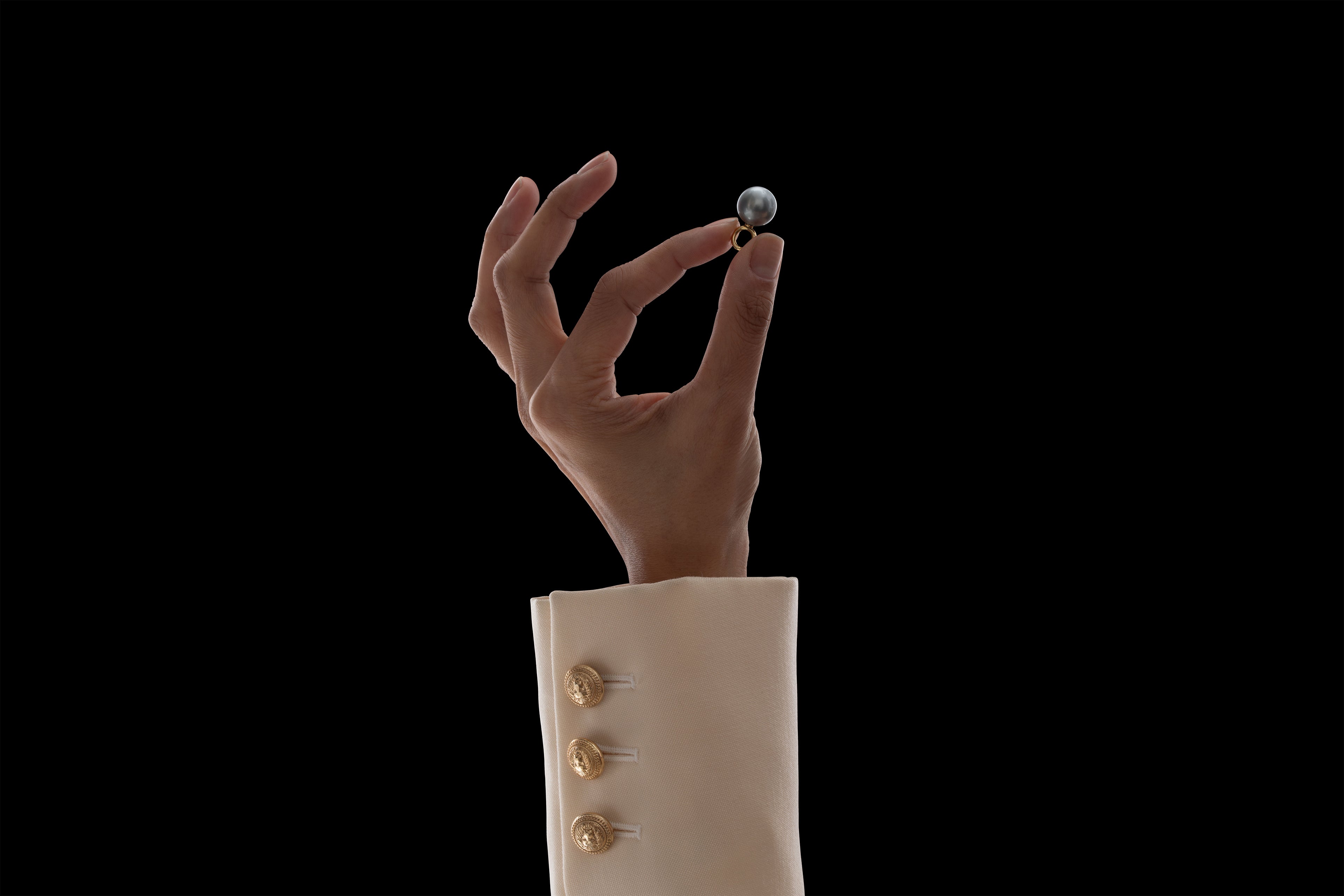 Ila's hand in a cream sleeve holding an ILA SODHANI Black Pearl Charm with a gold fitting in front of black background.