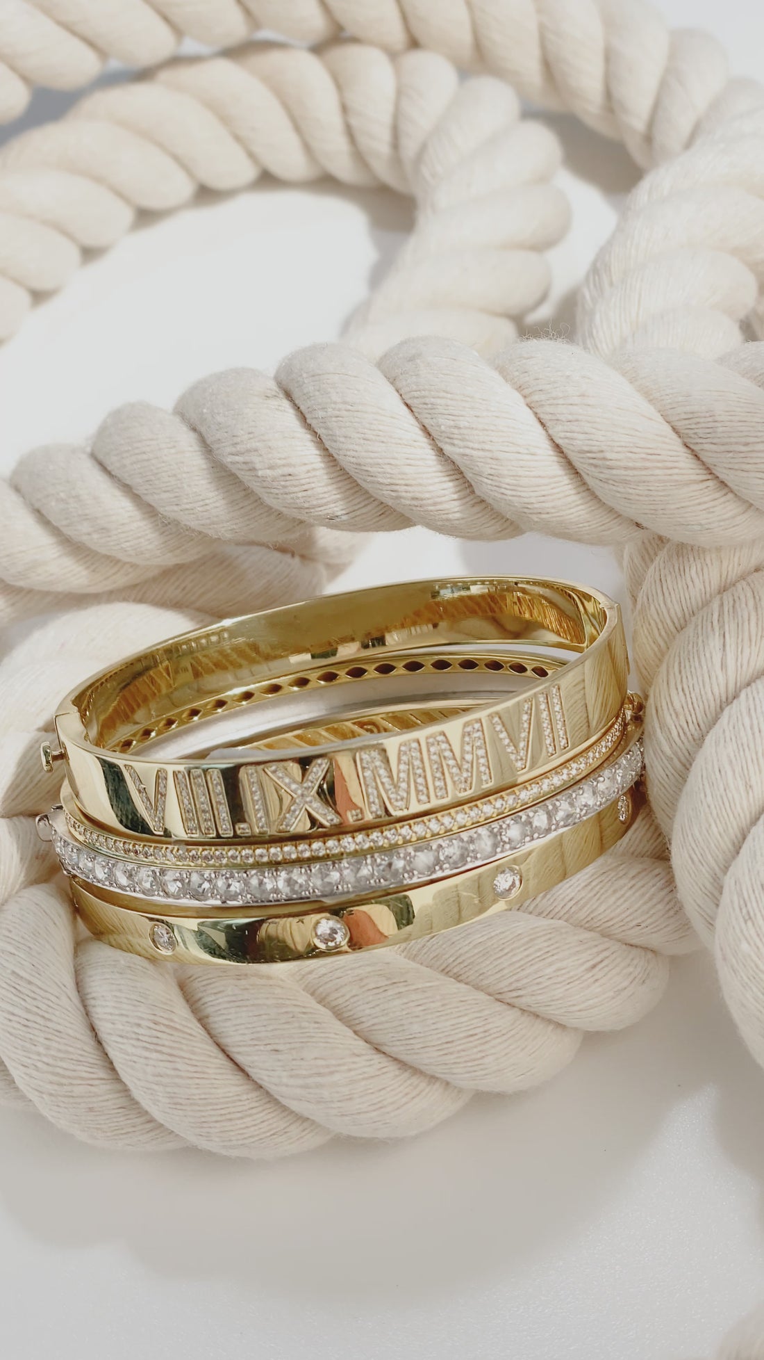 ILA SODHANI bangle stack shown on a coil of rope in front of a white background; from top to bottom: Celebration Bangle with natural diamonds in yellow gold, Aira bangle with natural diamonds in yellow gold, Winn Bangle with natural diamonds in white gold, Drake Bangle with natural diamonds in yellow gold.