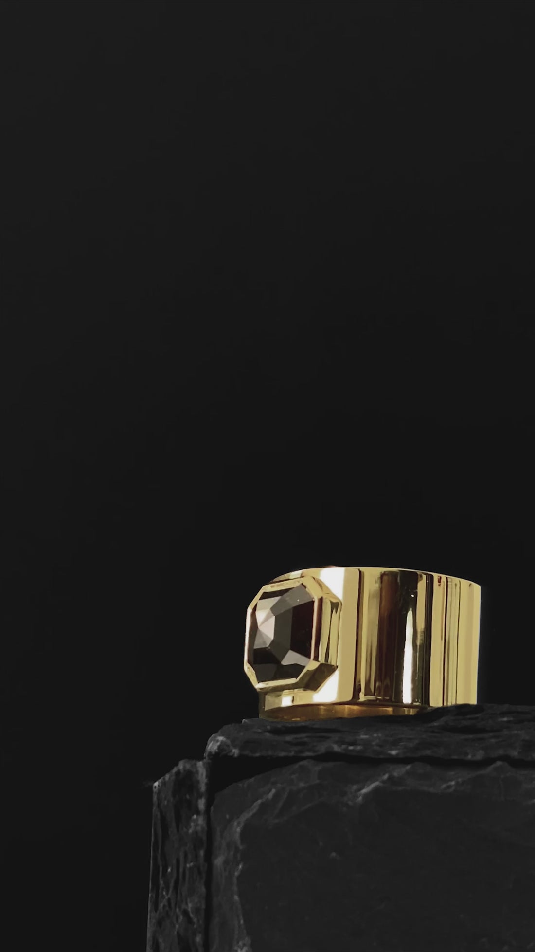 ILA SODHANI Aubergine Black Ring with a cushion cut black spinel stone set into a wide yellow gold band; shown on black stone in front of a black background with light moving across the ring.