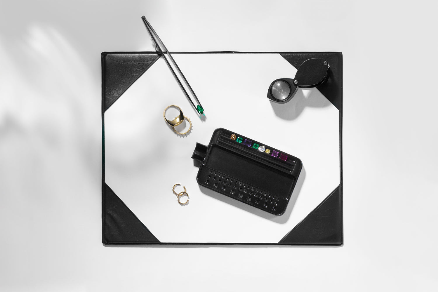 Ila Sodhani's desk with a black tray with sourced naturally colored gemstones with yellow gold pieces waiting to be set; an emerald is clasped in a pair of black tongs.